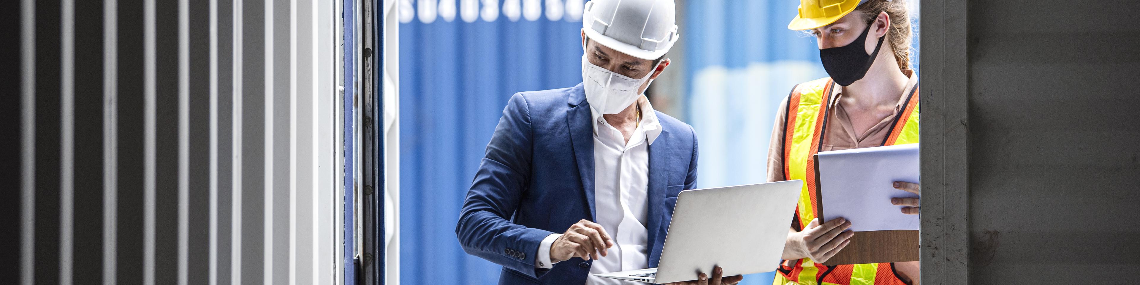 Business man and woman dock worker wearing safety protective clothing and face mask while working front of cargo container door open.