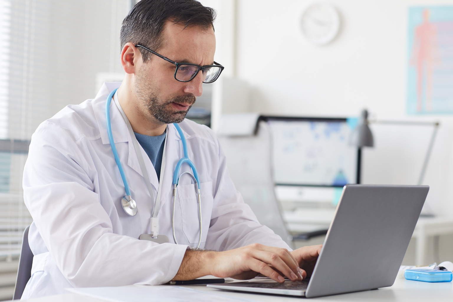 Serious male doctor in eyeglasses sitting at the table and typing on laptop computer in office