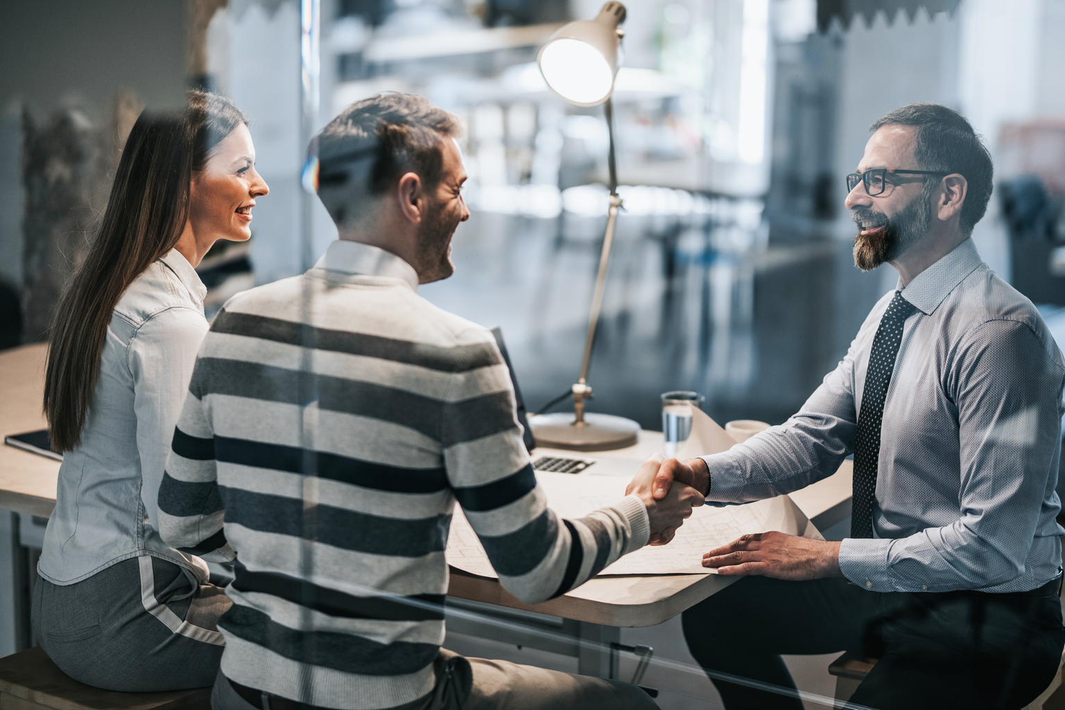 Happy real estate agent shaking hands with young couple after successful meeting in the office. The view is through glass.