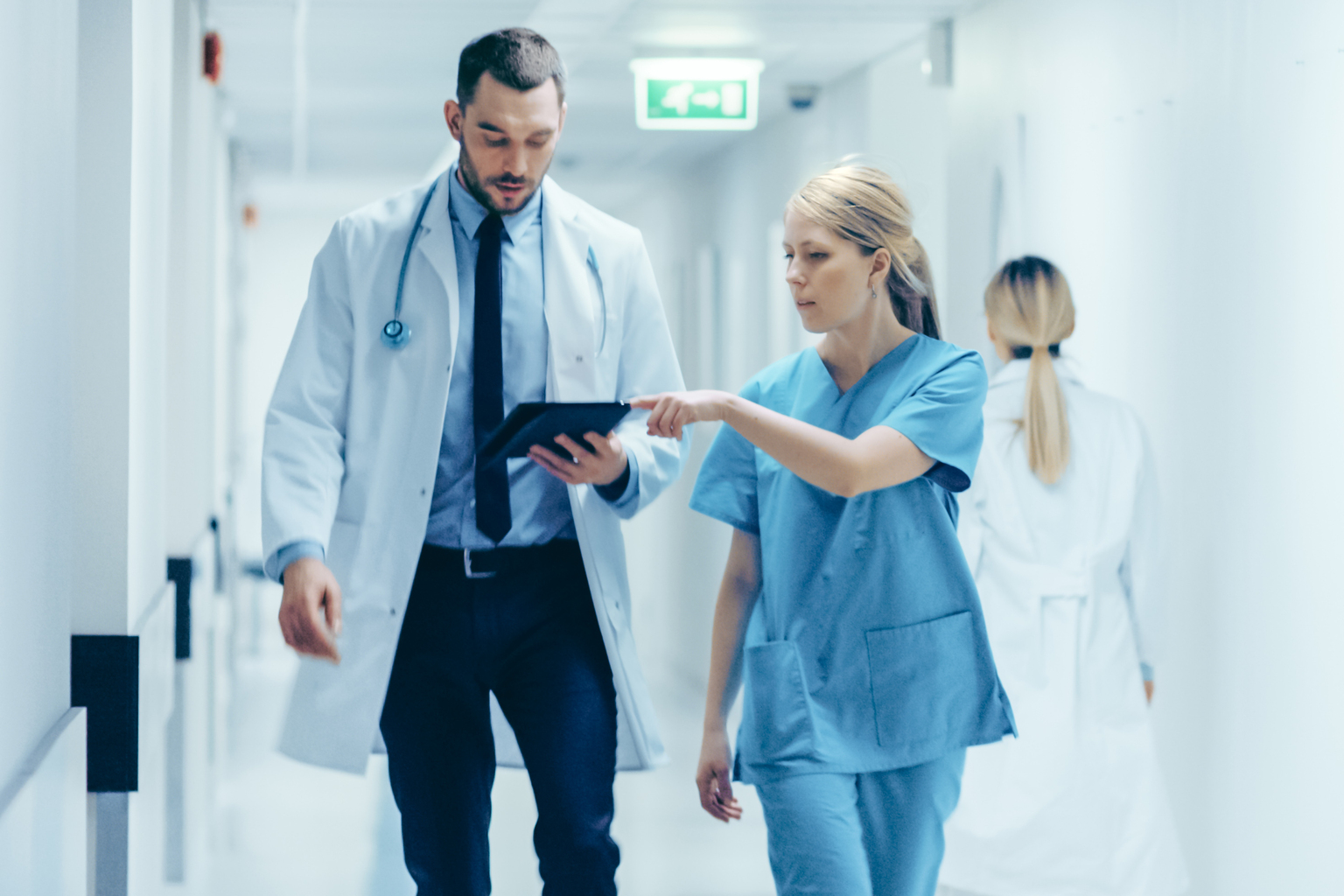 Female surgeon and male doctor walk through hospital hallway, consulting digital tablet computer while talking about patient's health