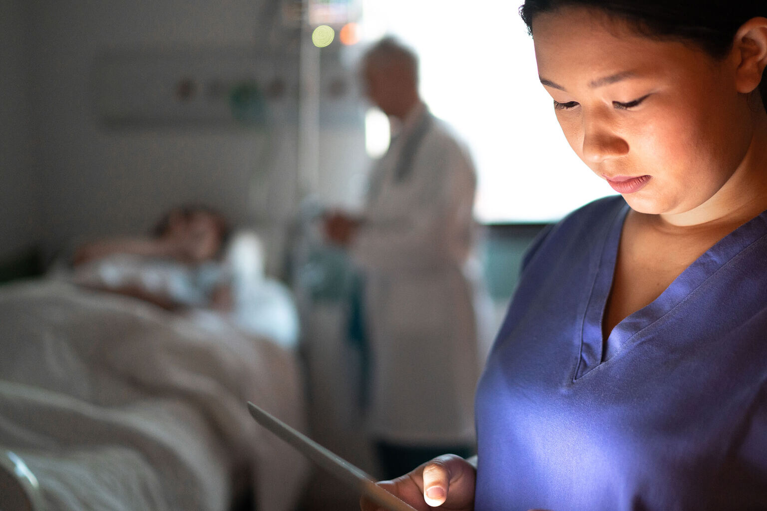 In dark hospital room, nurse reviews patient info on digital chart while doctor is at patient bedside in the background
