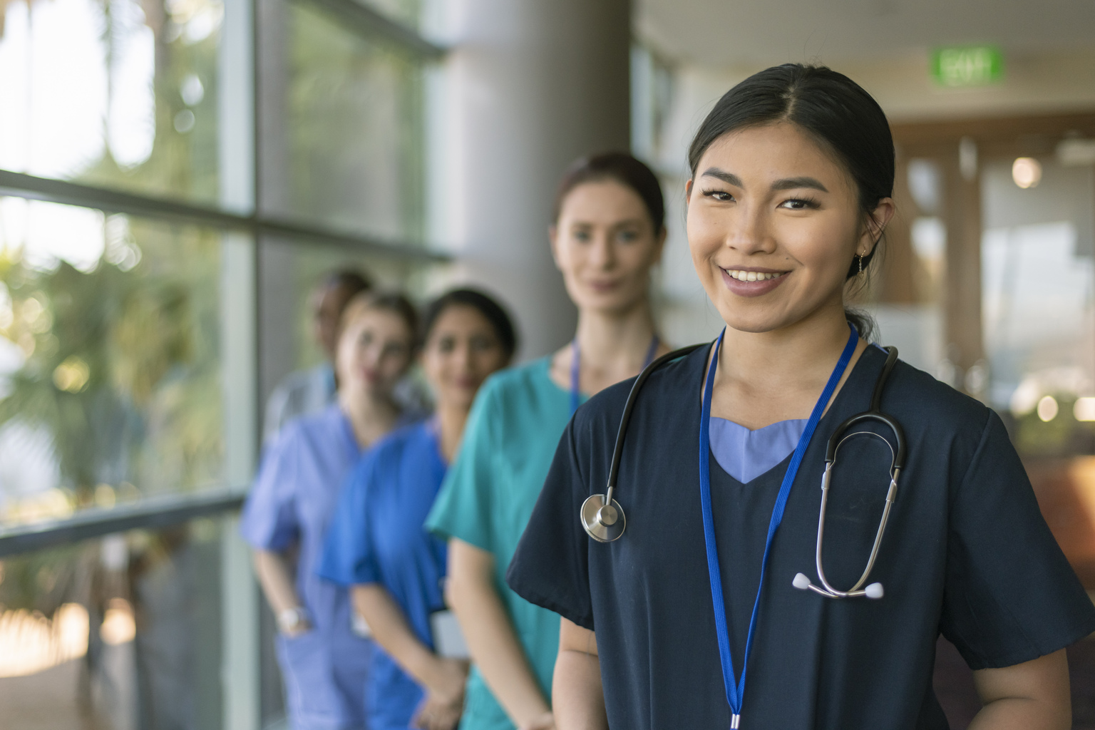 A female doctor of Asian descent poses with her multi-ethnic team of doctors and nurses and smiles directly at the camera while standing in a hospital corridor.