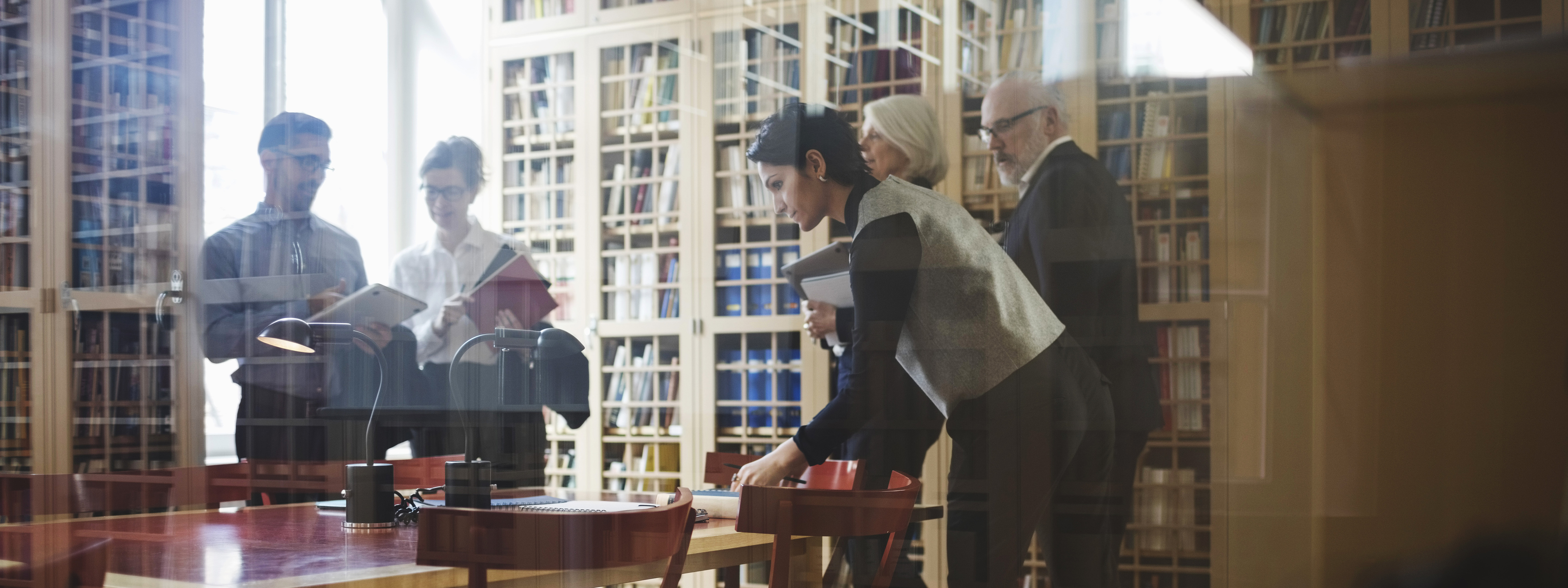 Five people in formalwear including active seniors to discuss a case near the bookshelf at law firm in Europe