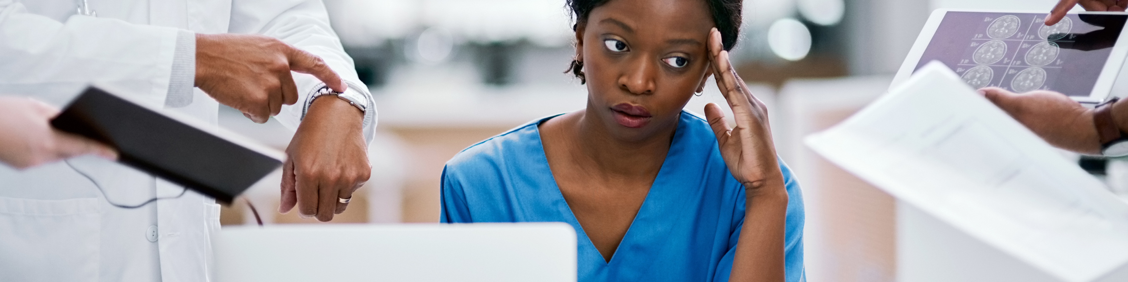 Burned out nurse sitting at table with laptop with multiple doctors around her requesting things