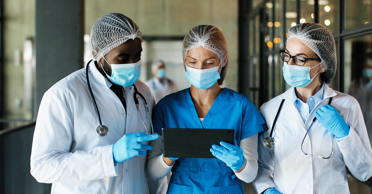 Mixed races male and females healthcare workers in hats medical masks and gloves walking in hospital and talking using tablet device