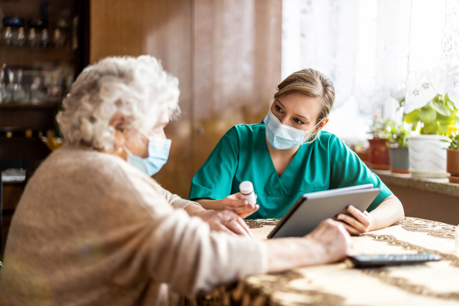Home nurse in facemask advising elderly patient on medication with tablet
