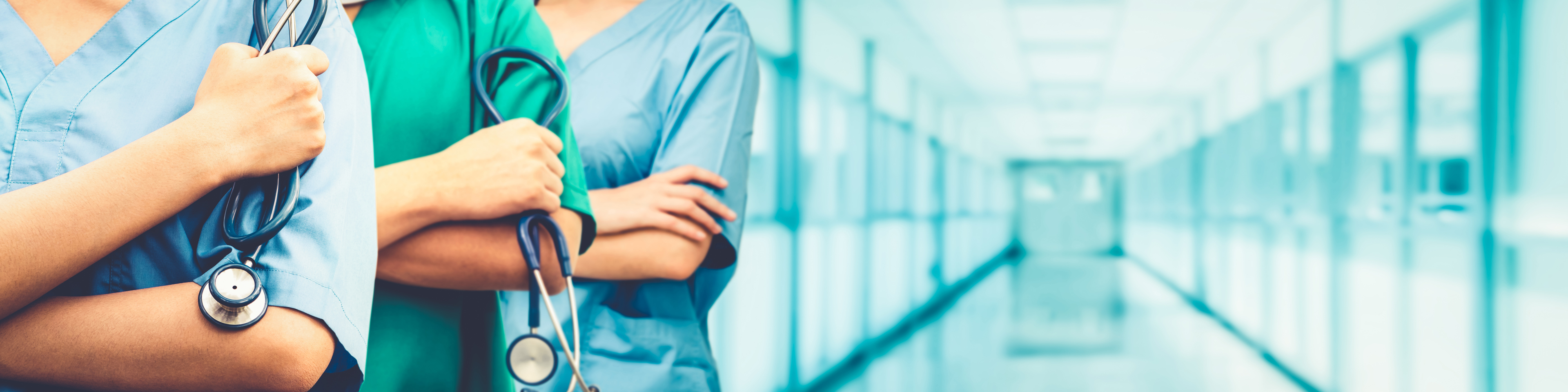 Three nurses lined up in a hospital hallway with their arms crossed, two holding stethoscopes