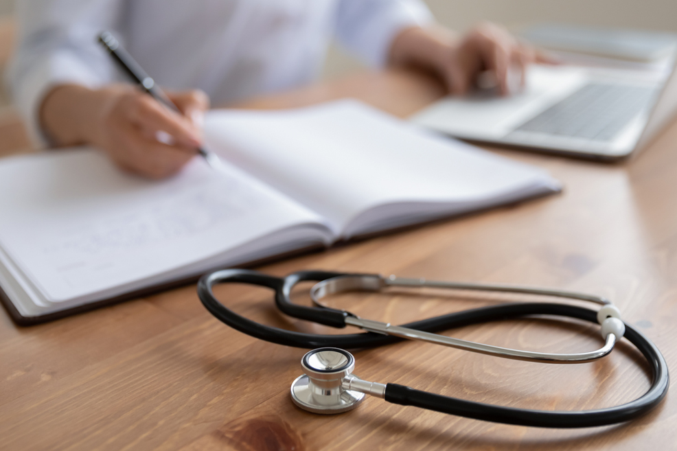 Female doctor sitting at desk in front of laptop writing in a medical journal, stethoscope in foreground on desk