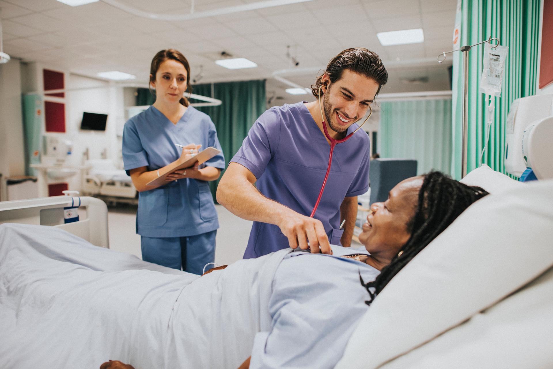 Young male nurse checking female Black patient with stethoscope in hospital, nursing student observing