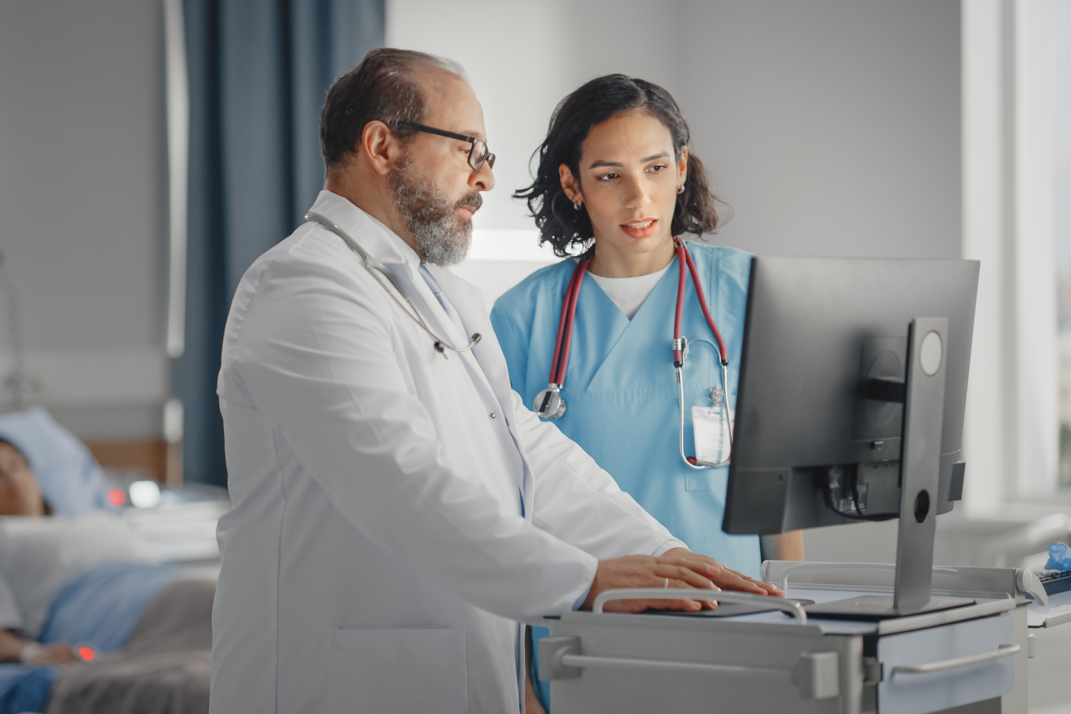 Doctor talks with head nurse at computer with patient in hospital bed in the background