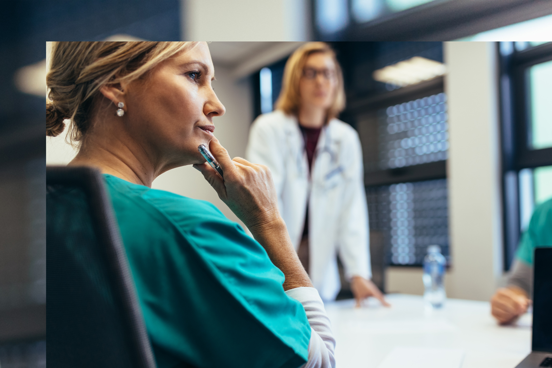 Focus on nurse sitting at meeting room table with doctor standing, leaning on table in background