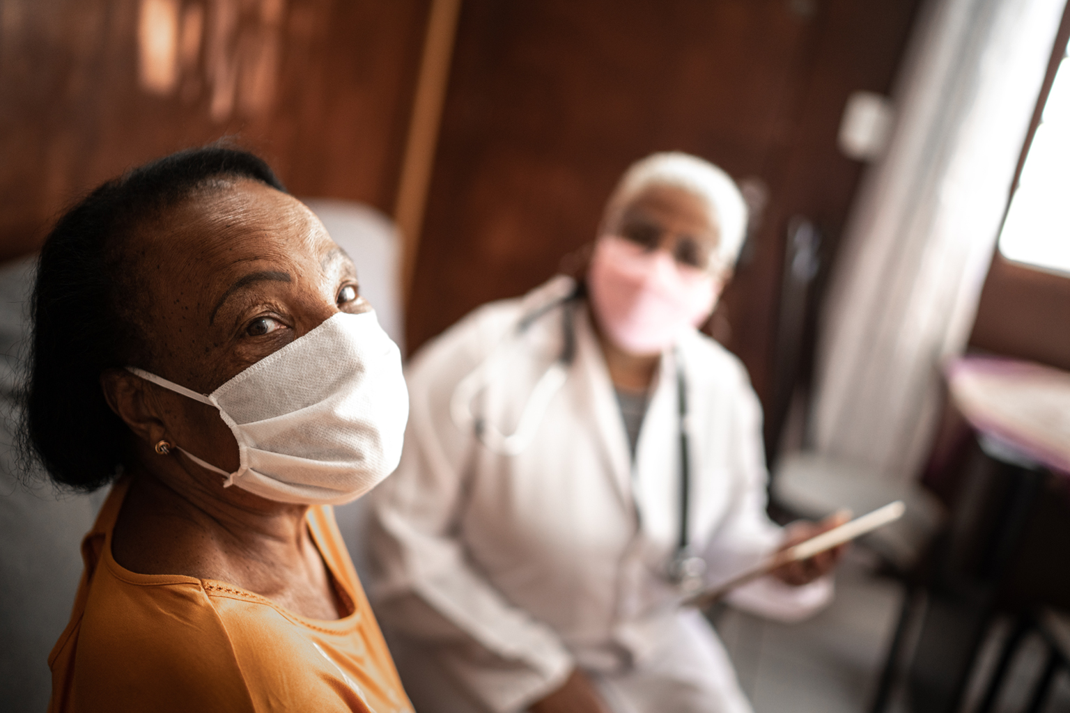 Older woman sitting in office with doctor, both wearing masks