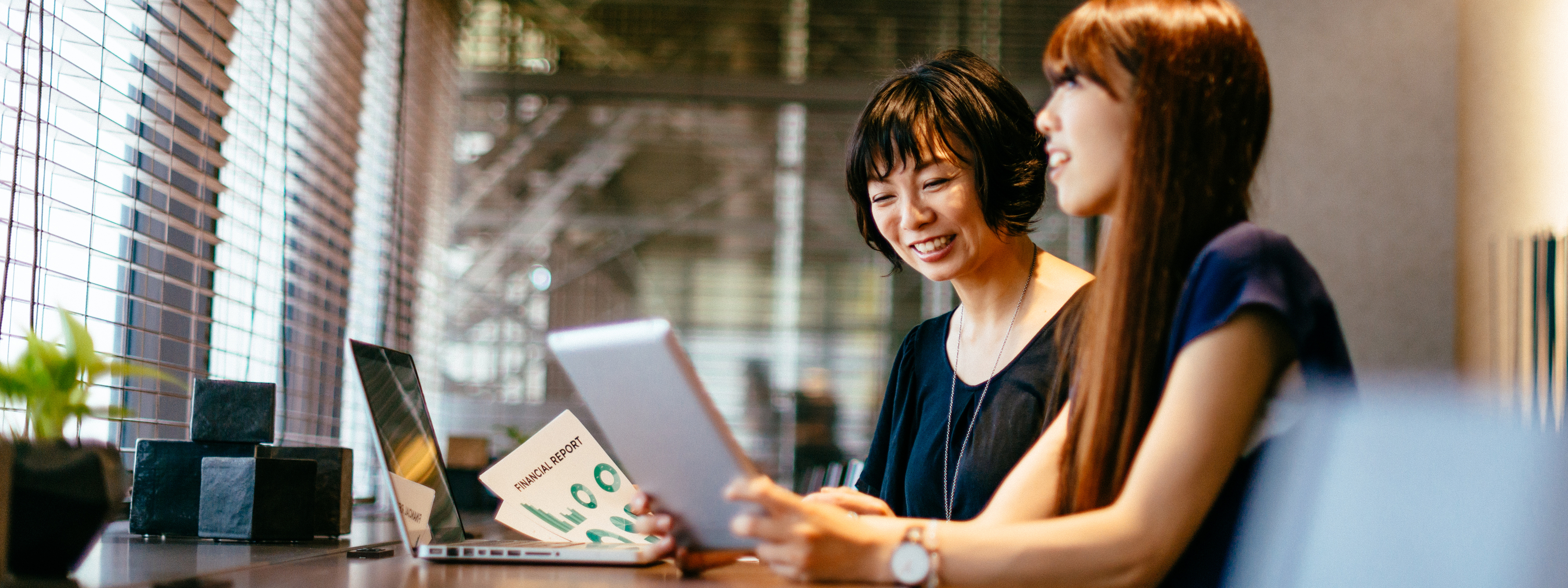 Asian female professionals reviewing financial reports on tablet and laptop.