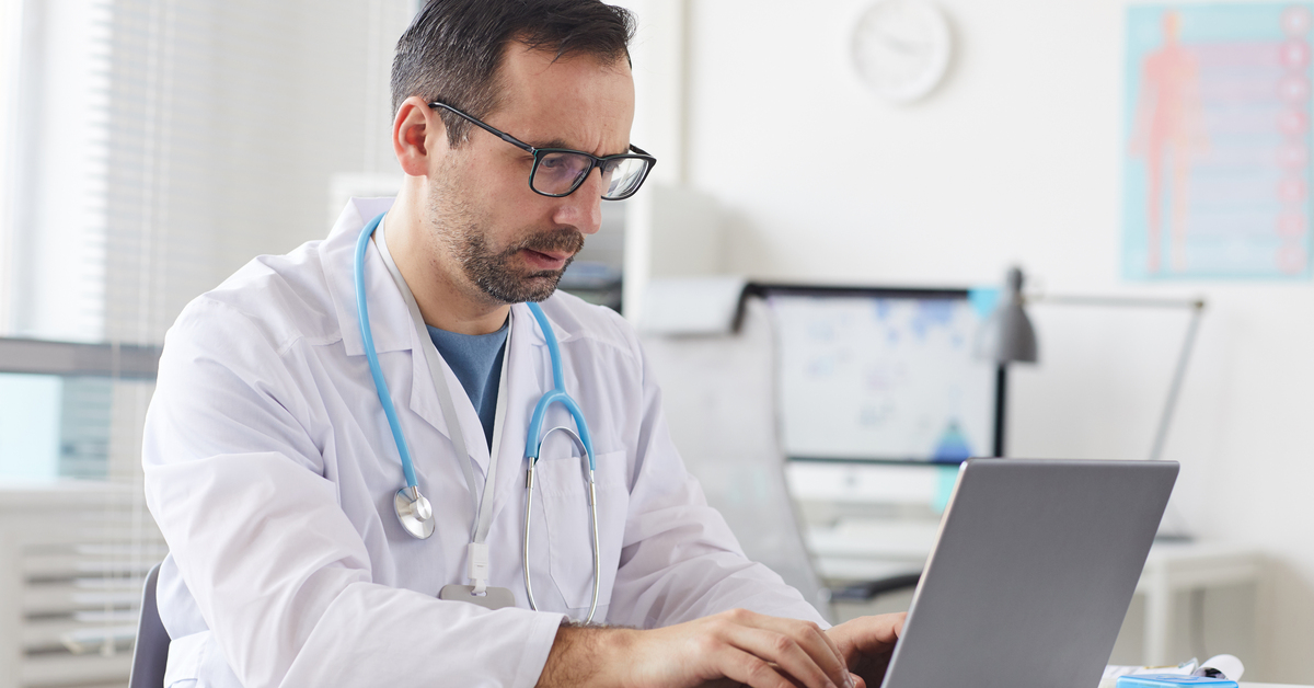 Serious male doctor in eyeglasses sitting at the table and typing on laptop computer in office