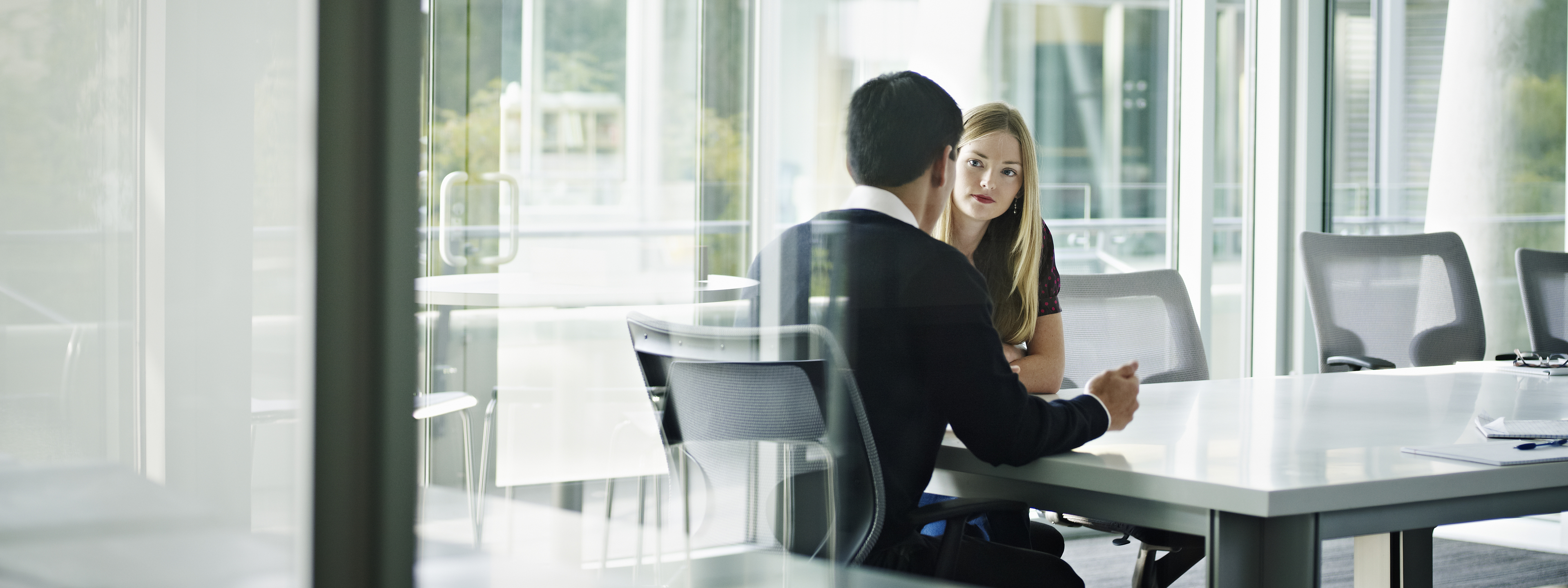 Businesswoman and businessman in discussion at table in glass walled conference room.