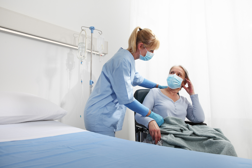 Nurse helping patient in chair in hospital room, both wearing masks