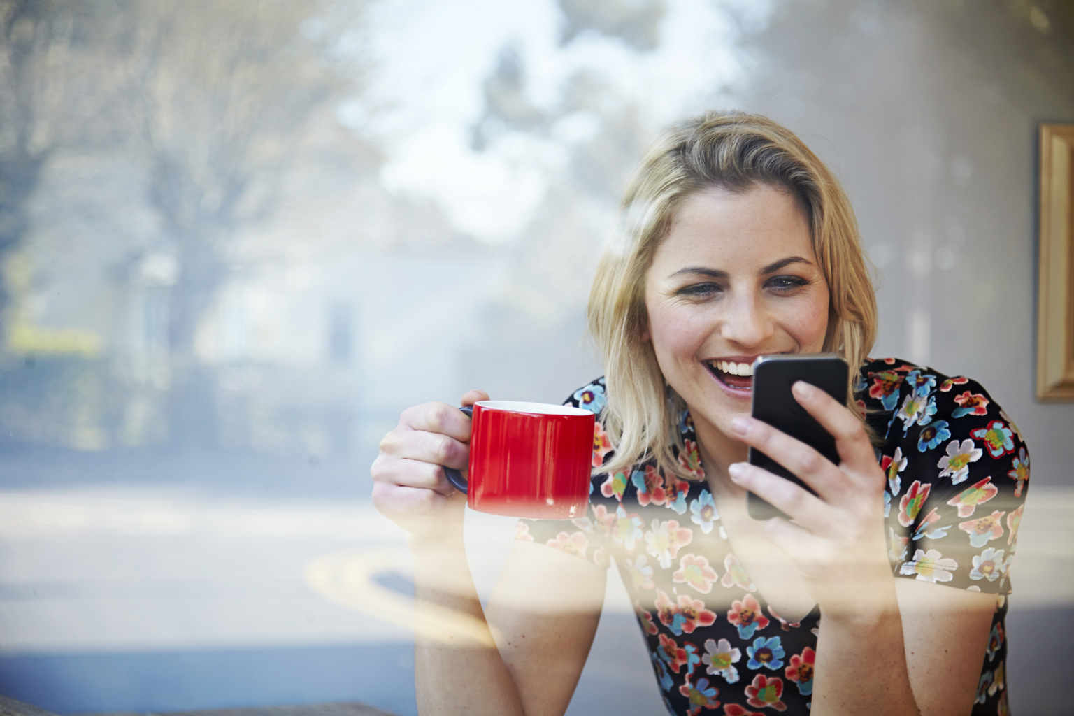 Woman laughing at her phone in coffee shop window.