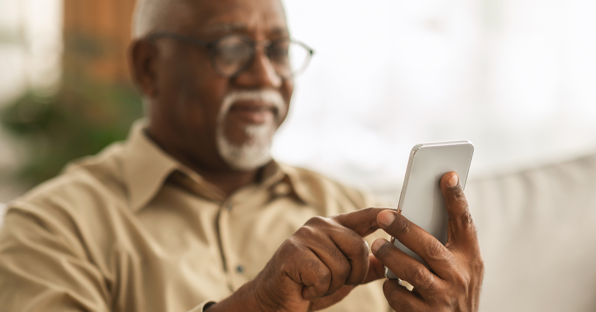 Senior African Man Using Smartphone Texting Sitting On Sofa Indoor