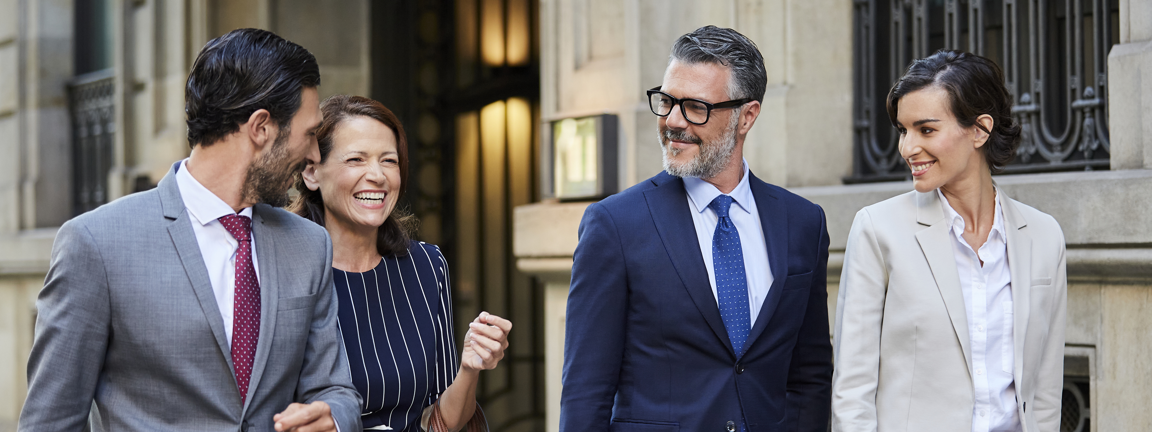 Smiling businessmen and businesswoman looking at female colleague laughing. Professionals are walking together outside hotel. They are in businesswear.