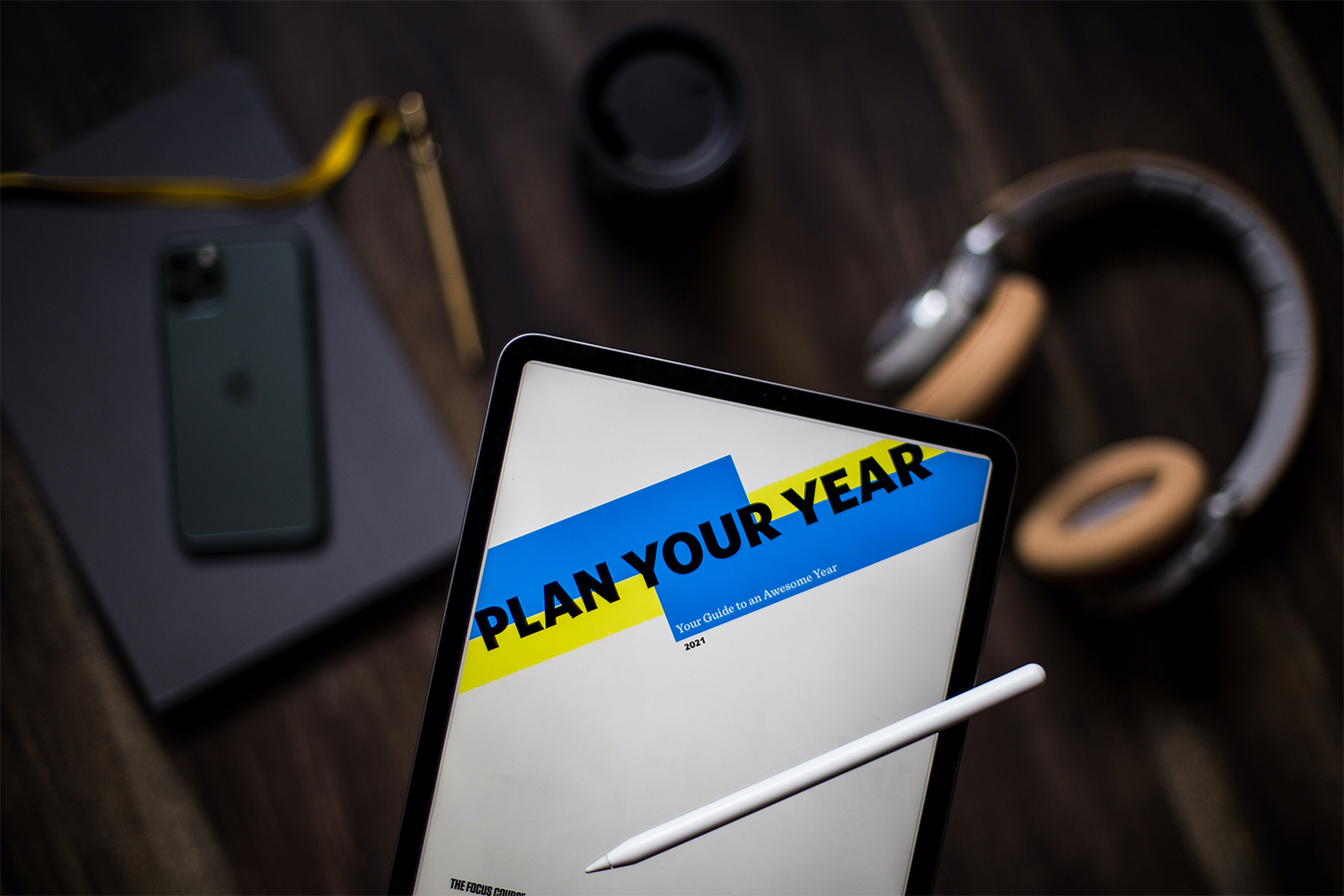 Plan your year 2021 on a tablet with a tablet pen in foreground; phone, laptop, travel mug, and headphones in background
