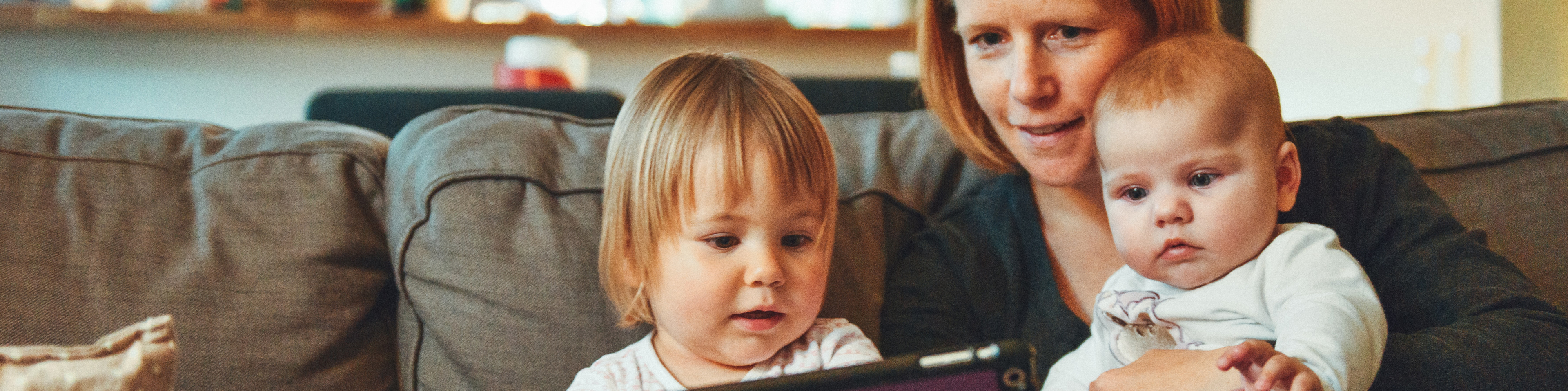 Woman sitting on couch with two young children, all watching tablet