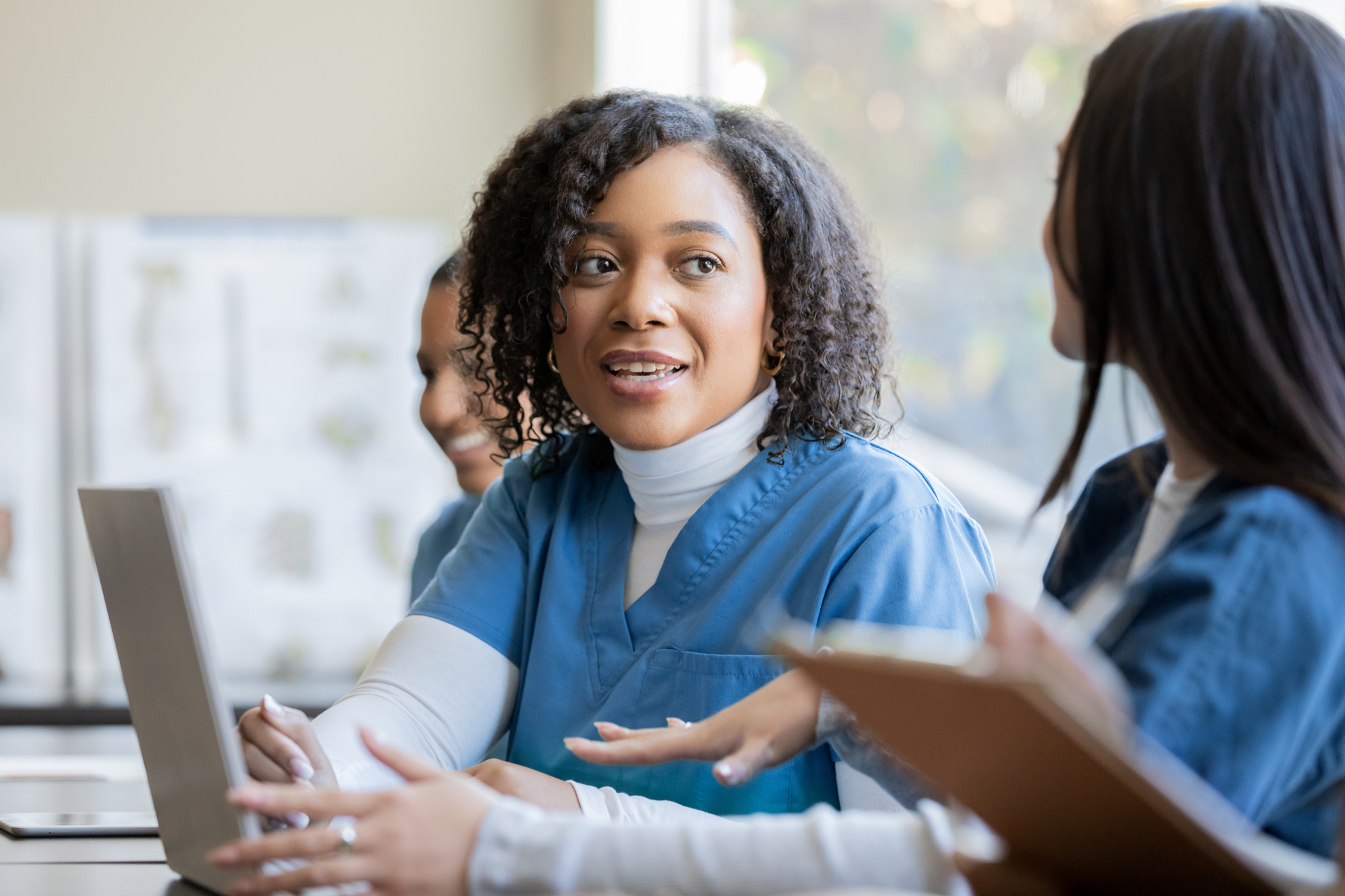 A nursing student wearing a white turtleneck under blue scrubs chats casually to a classmate sitting next to her