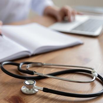 Female doctor sitting at desk in front of laptop writing in a medical journal, stethoscope in foreground on desk