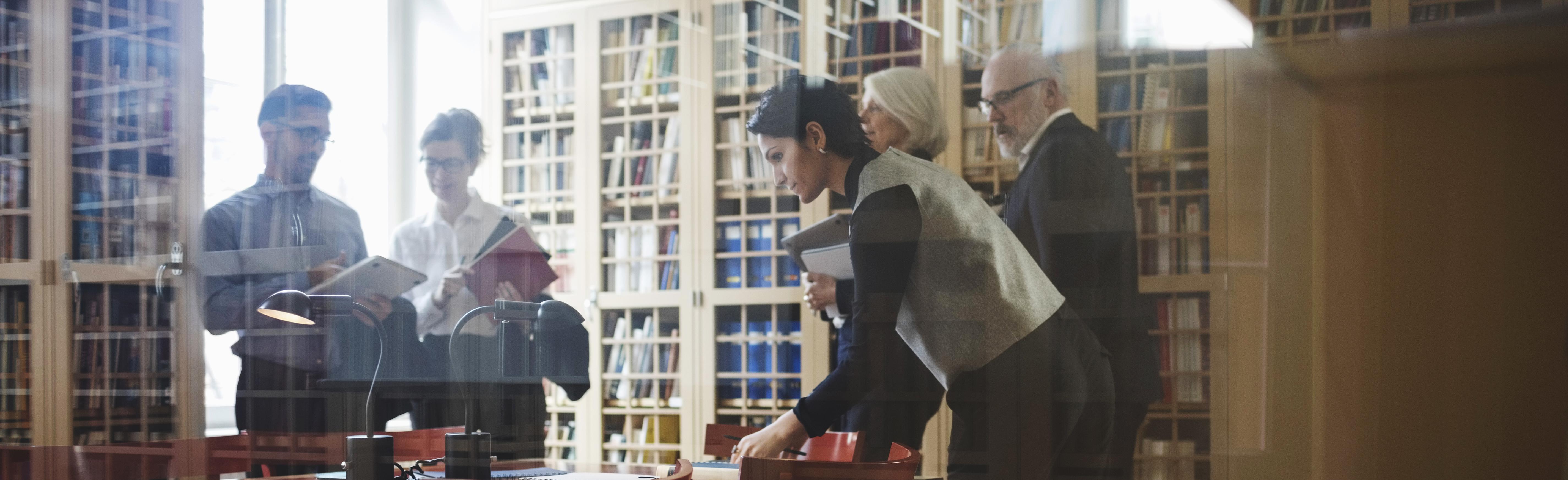 Five people in formalwear including active seniors to discuss a case near the bookshelf at law firm in Europe