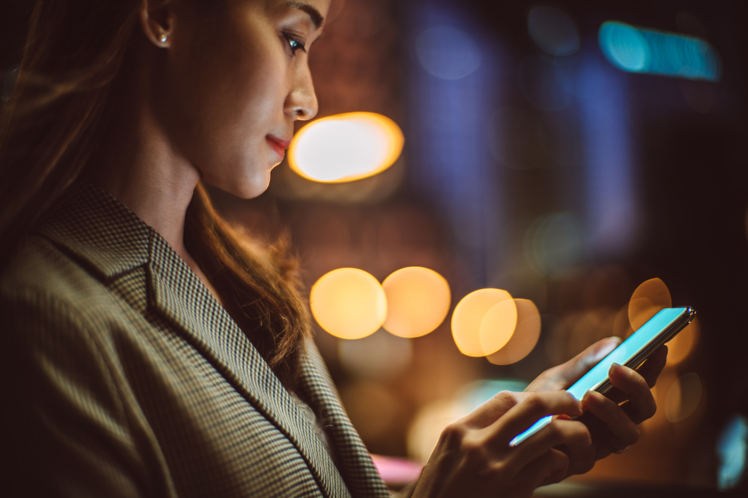 Young Asian woman using smartphone in downtown district, against illuminated city buildings