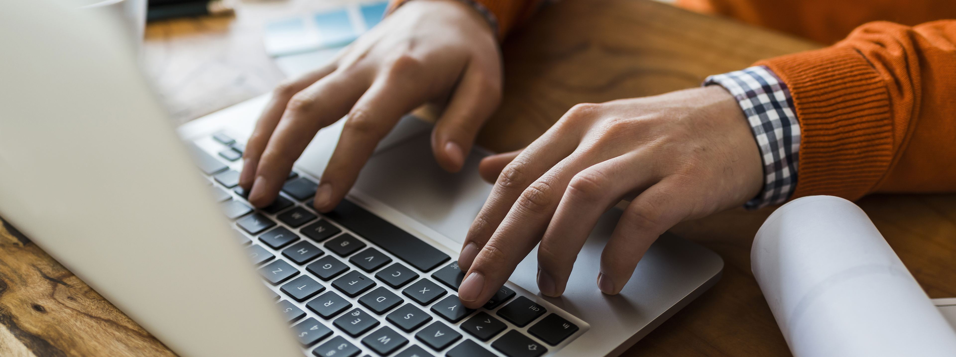 Man with orange sweatshirt, and flannel sitting at a wood table, typing on macbook with architectural plans on desk, Q3 2021, TAA NA US - Preparer