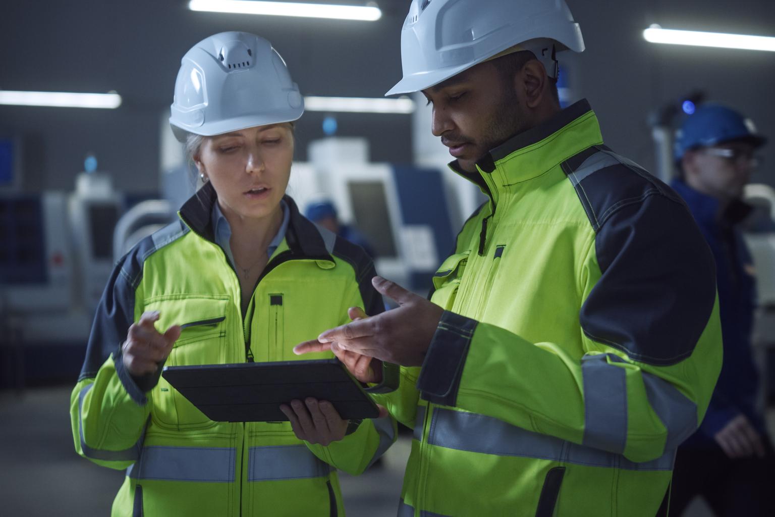 Chief Engineer and Project Manager Wearing Safety Vests and Hard Hats, Use Digital Tablet Computer in Modern Factory, Talking, Optimizing Production Line. Industrial Facility with CNC Machinery