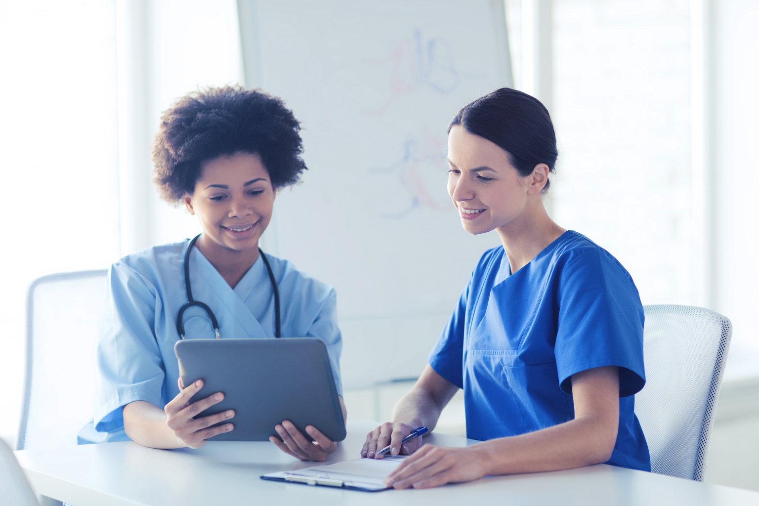 Two female healthcare professionals dressed in scrubs working on a tablet together