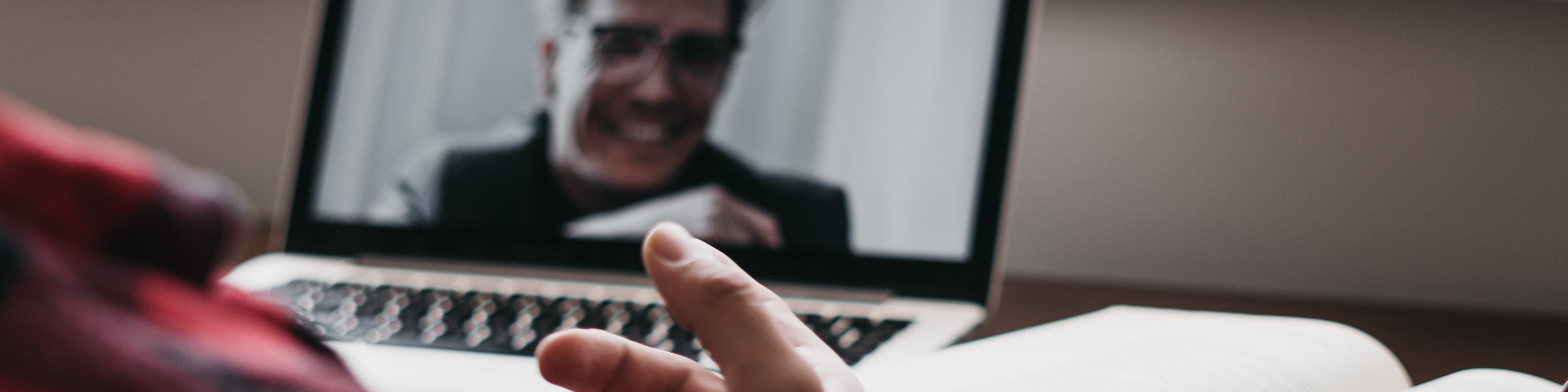 Close up view over man's shoulder looking at laptop and book on table, another person on laptop screen