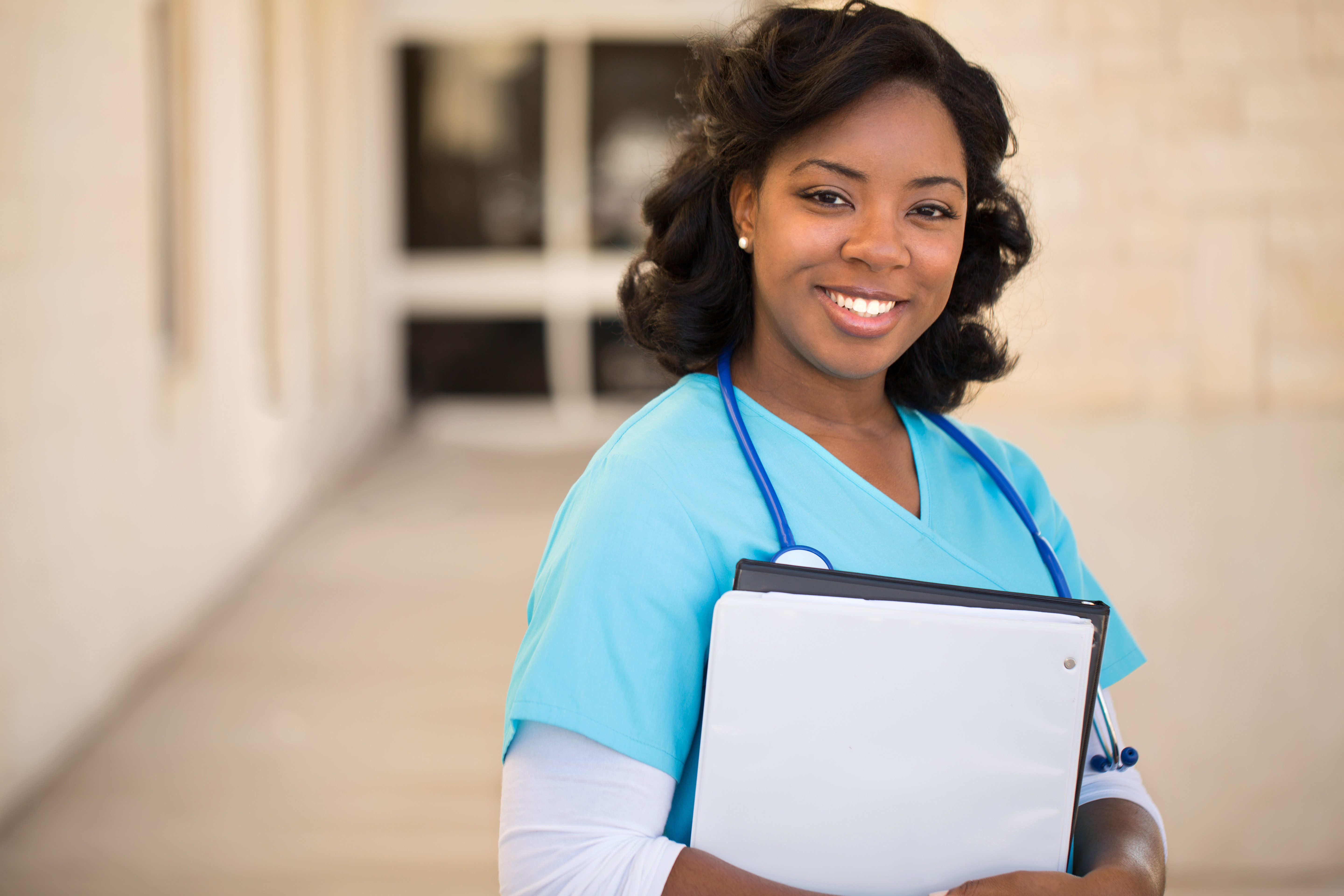 Nurse holding binder of documents smiling at the camera, wearing light blue scrubs. 