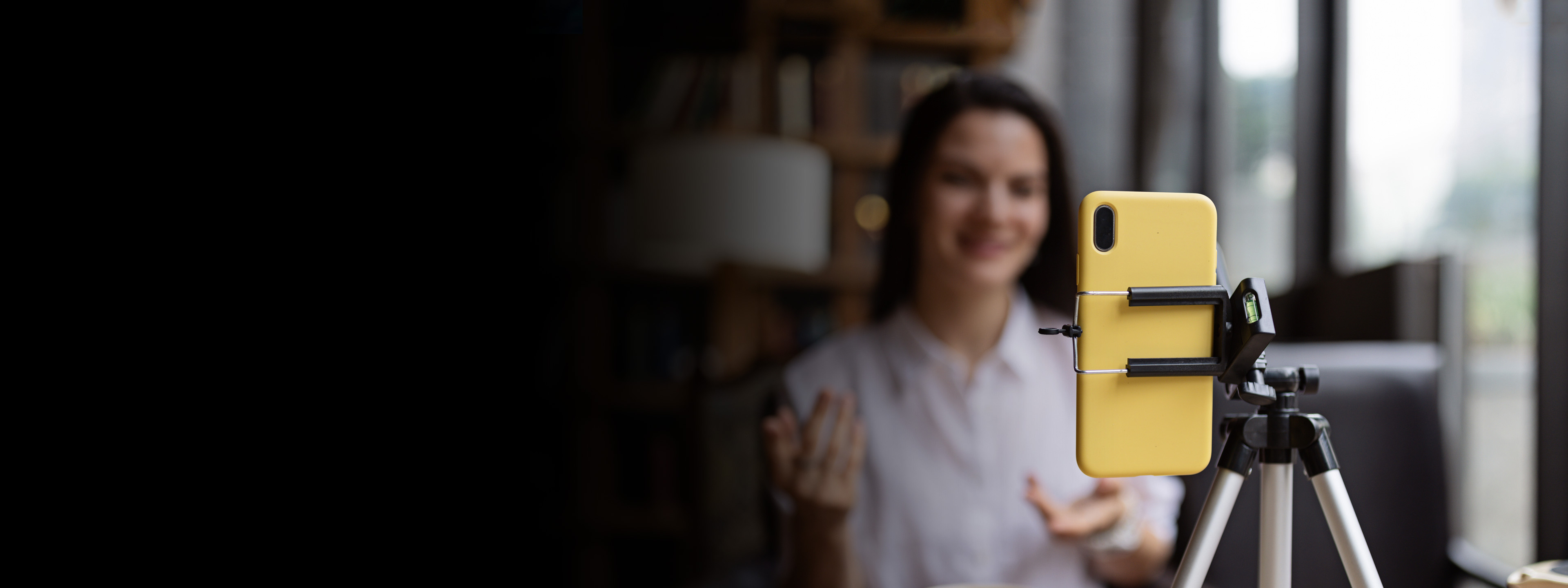 Happy young Caucasian millennial or gen z woman with long brunette hair streaming with smart phone on tripod, shooting social media blog in modern cafe.