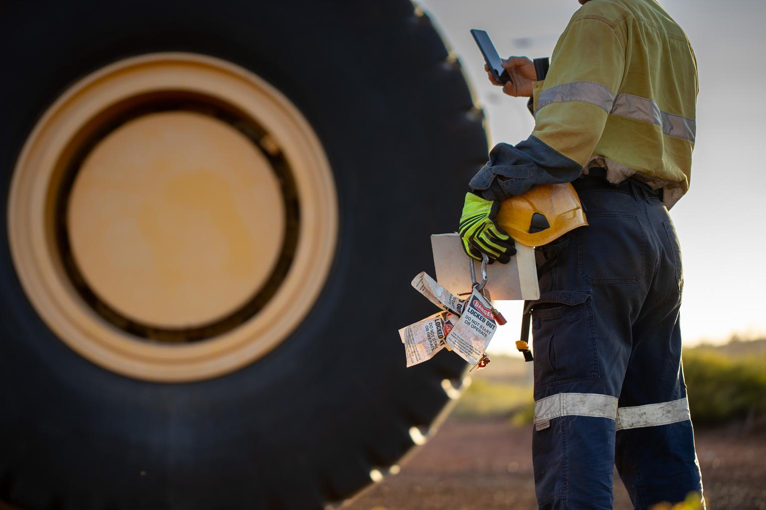 Haul truck inspector wearing work uniform safety glove holding hard hat danger tags personal locks inspection pre operation book inspecting taking pic with cell phone defocused haul truck background
