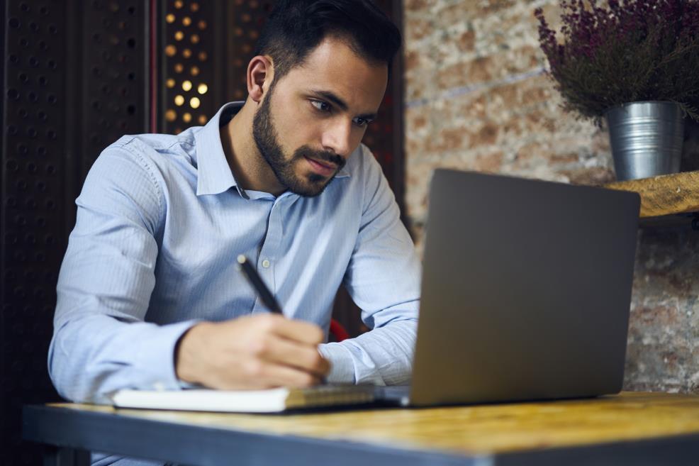 Man sitting at small table with laptop, notebook and pen, watching something on the screen