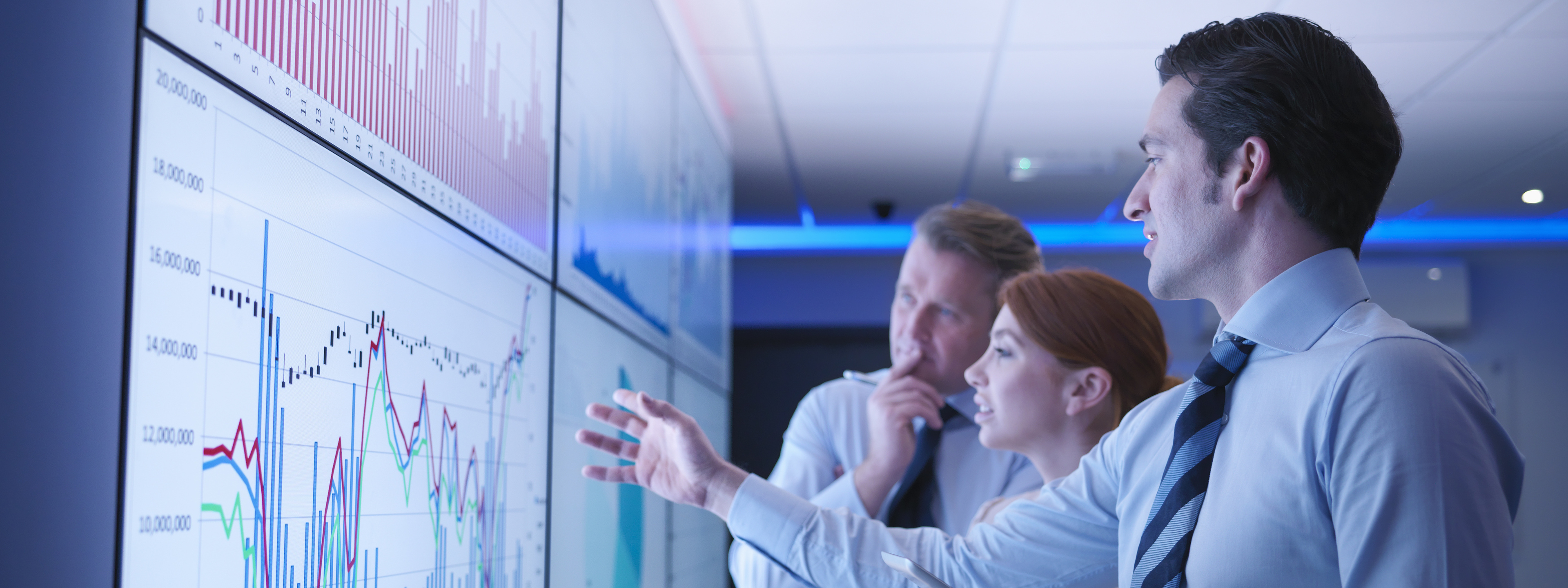 Three business people discuss graphs on screen in meeting room