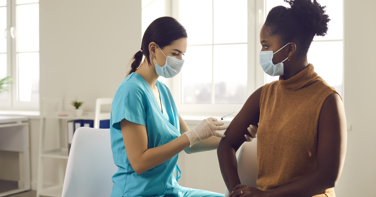 Young African American woman in medical face mask sitting at doctor's office and getting a shot in her arm, nurse in medical face mask holding syringe and giving female patient modern covid 19 vaccine injection