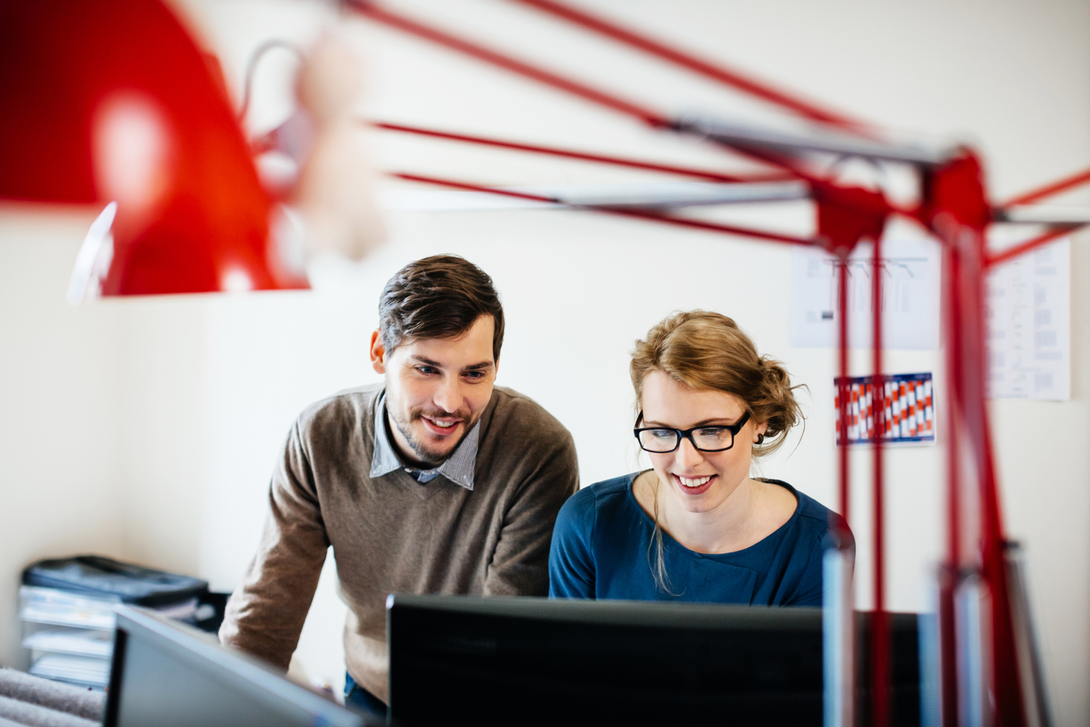 Two young casual start up business people standing in their office behind a desk and looking at a computer monitor. Both are smiling