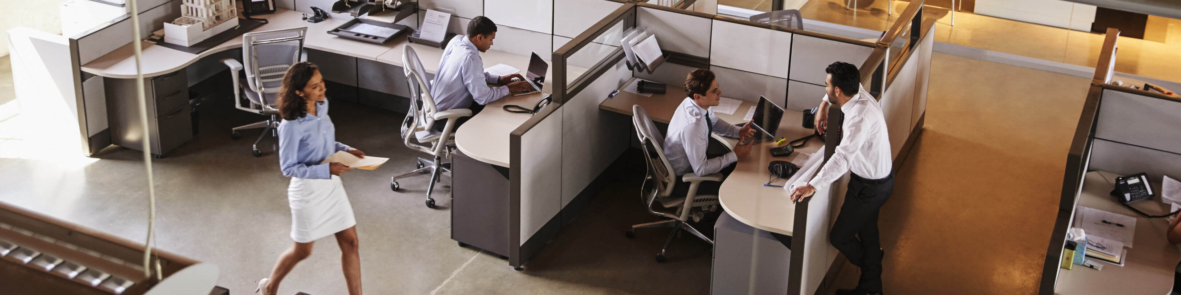 overhead view of a cubicle farm with walls at half-height; two people sitting at desks with computers, one man leaning over the top of one cube wall to talk to another, a woman walking past another cubicle with papers in her hand.