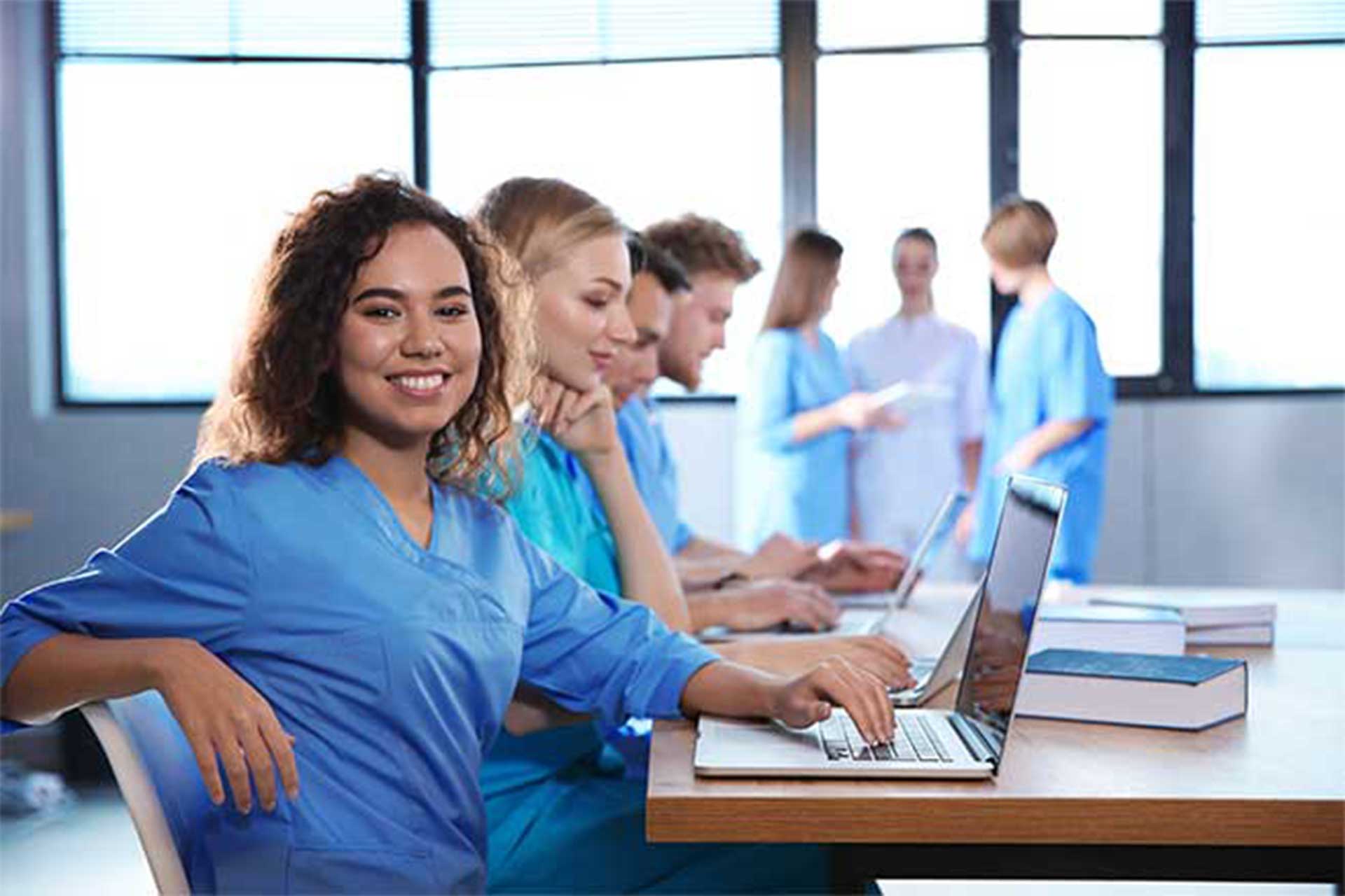 Group of nursing students in meeting room, some chatting, some on laptops, woman in front smiling at camera