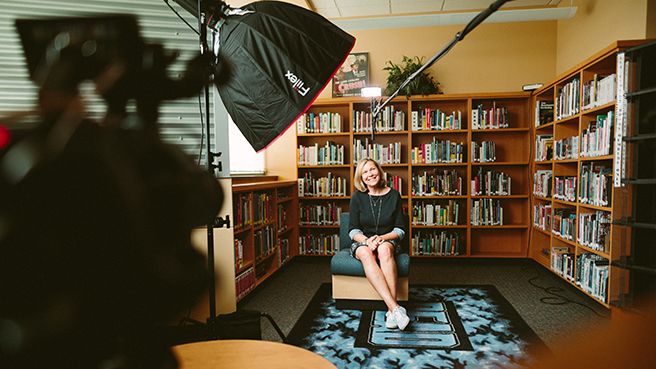 Woman sitting in a library for an interview, image taken from behind equipment including camera