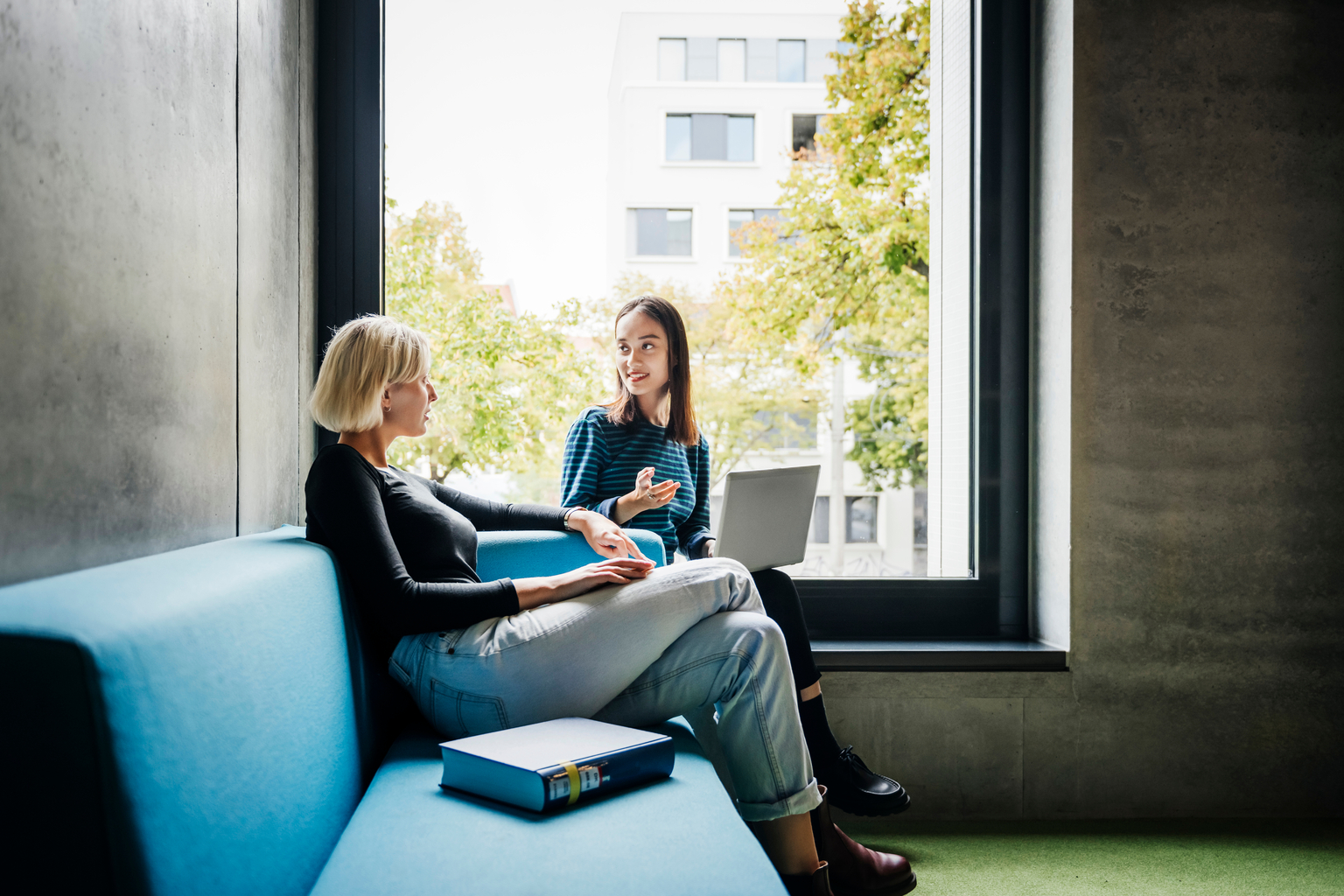 Two friends chatting by a large window in a quiet library space together.