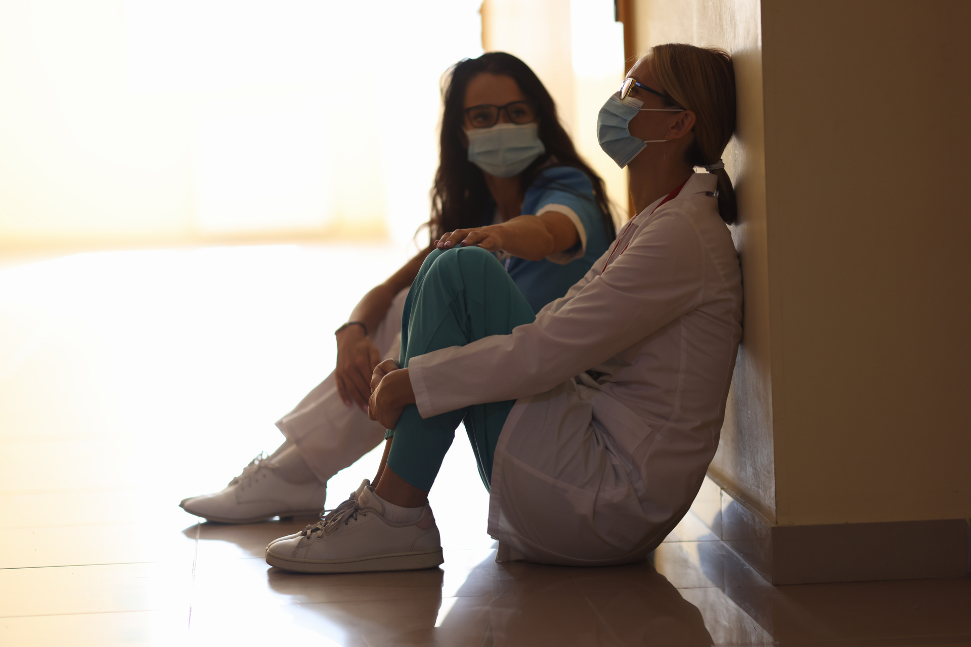 Nurse wearing face mask, slumped against wall in frustration; nurse colleague comforts her by placing hand on knee