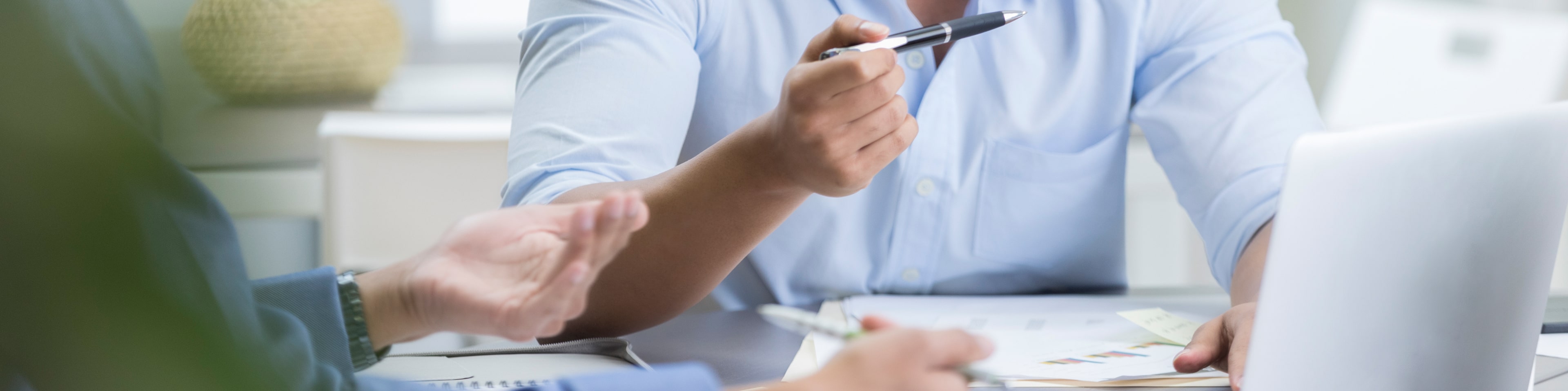 Young businessman has a serious discussion with male colleague or client. An open laptop is on the desk. He is gesturing with a pen.