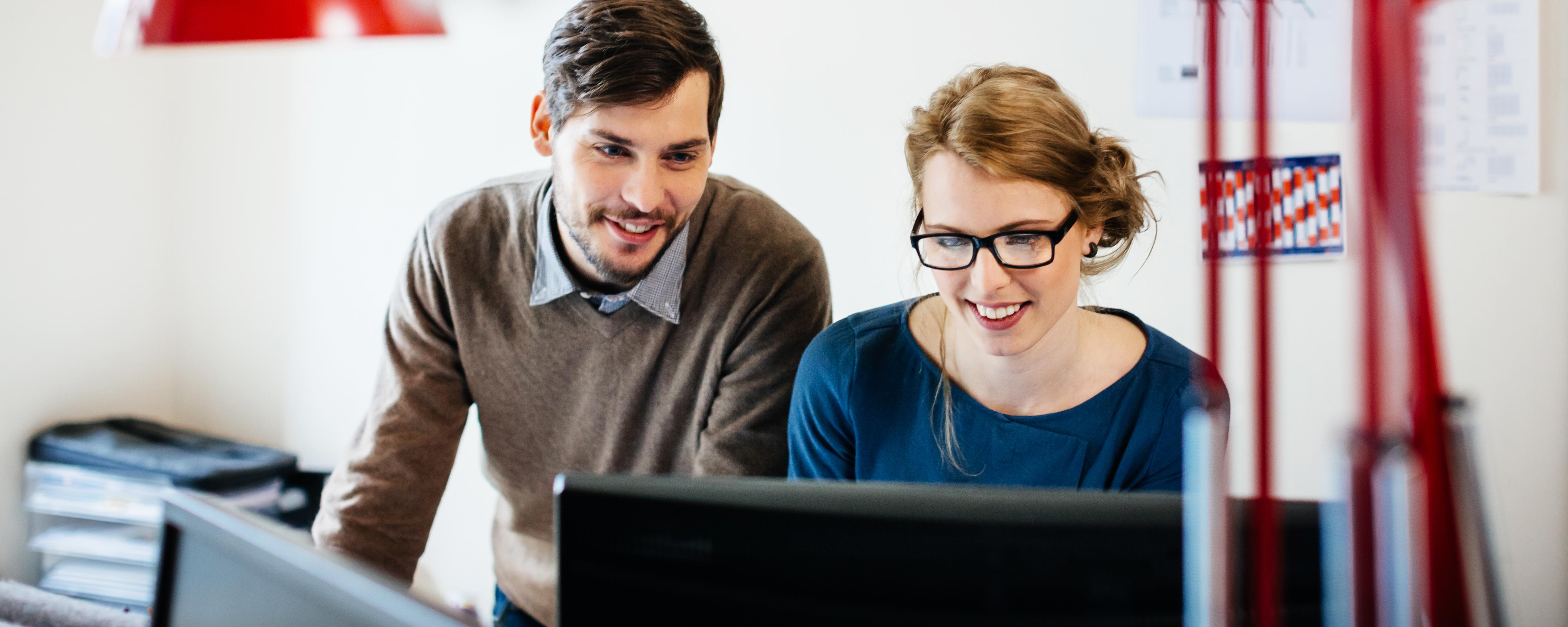 Two young casual start up business people standing in their office behind a desk and looking at a computer monitor. Both are smiling