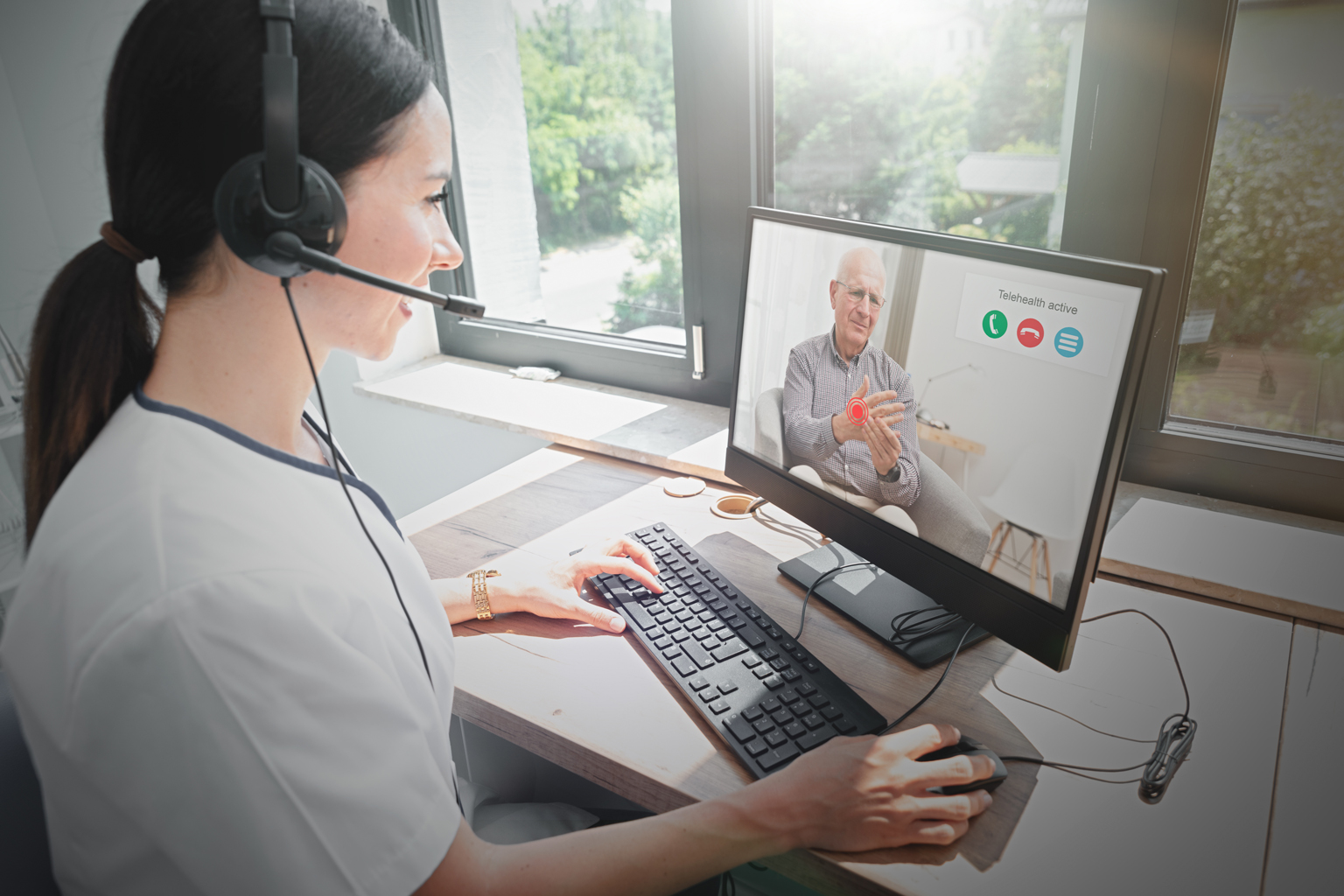 Healthcare provider sitting at computer with headset, talking with patient on screen