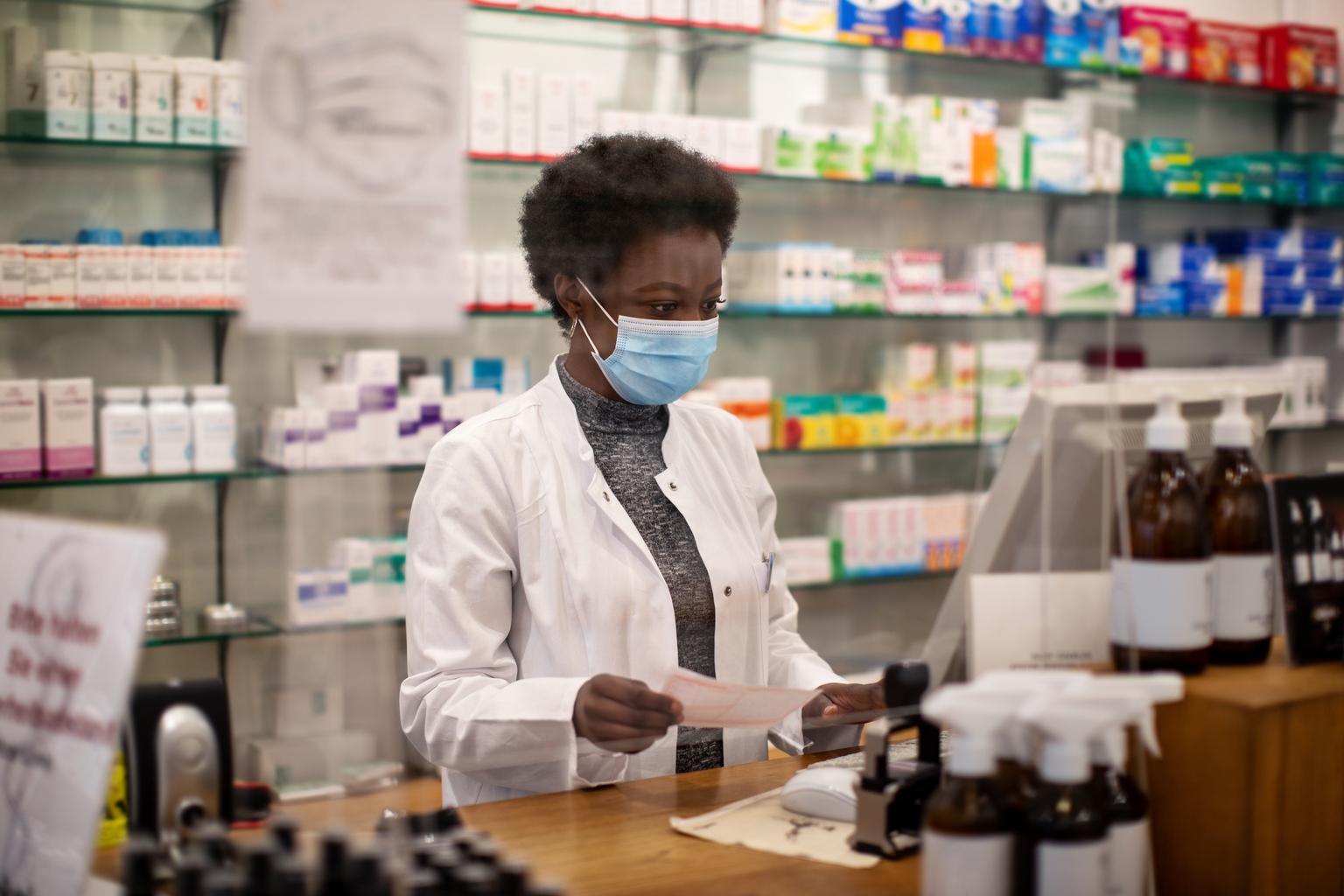 Female African pharmacist wearing surgical face mask checking out customer's medication prescription. 