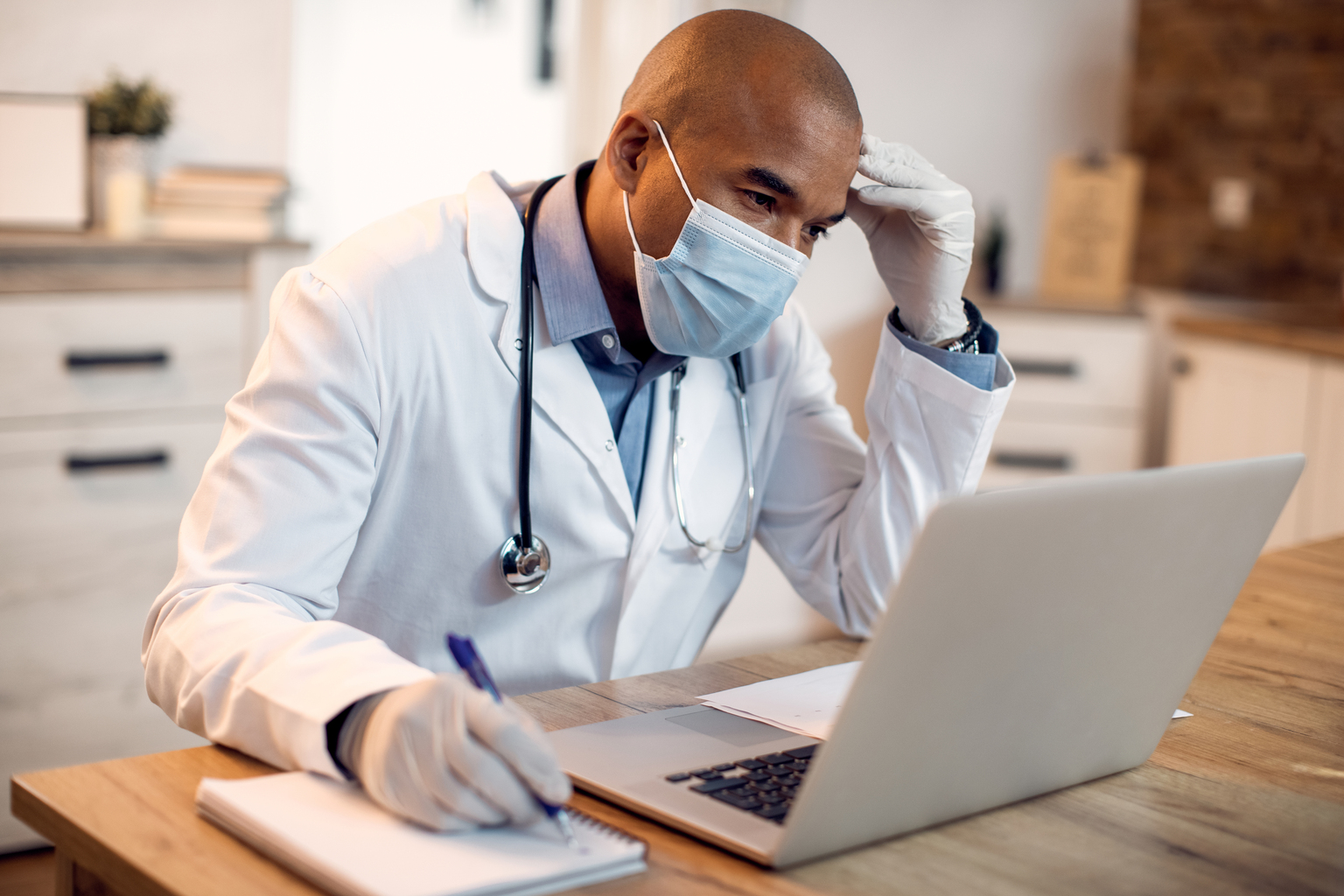 Pensive Black doctor with mask taking notes while working on laptop at his office during pandemic